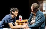 Side on picture of Colin Morgan and Roger Allam sat at two connecting sides of a table, their chairs are pulled round so there's no table between them. Colin, in the polo shirt, is leaning over towards Roger (in shirt and blue knitted cardigan). Their hands are entwined on Roger's lap. Roger looks confused and Colin like he's explaining something.