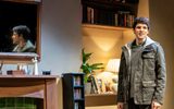 We see the back wall of a living room, a wooden hearth with various trinkets on top like photos in frames and decorative figures, a CD rack filled with CD's and shelves filled with books and a plant and lampshade. To the right of the scene stands Colin Morgan, this time in a green knitted jumper, unzipped grey hoodie and outdoor jacket on top. He looks agitated.