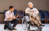 A low shot, in the blurred background are hospital chairs lined against the back wall. In the middle of the stage is Simon Williams in a wheelchair with a tartan blanket over his legs, he has headphones round his neck. He's looking at Sacha Dhawan who is on his knee next to him and reading a book to him.