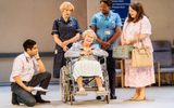 Production photo from Allelujah! The scene visibly a hospital with beige walls and blue signs listing different departments plus faded old chairs. In the middle of the stage is Jacqueline Clarke in a wheelchair. Crouched on the left is Sacha Dhawan in a suit with a stethoscope around his neck. Behind the wheelchair is Deborah Findlay in a dark blue medical smock dress, and next to her is Nicola Hughes in a light blue nurse shirt and dark scrub trousers. On the right is Rosie Ede with neat brown hair and a pink floral mid length dress, she is smiling at Jacqueline.