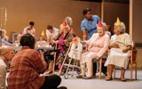 A low angle shot of the stage. The care home residents are sat in various formations, on plastic chairs or larger cushioned ones. They're all wearing colourful pointy party hats in reds, yellows and oranges. One woman has a zimmer frame with a sunflower attached to it. In the foreground out of focus is Nadine Higgin facing the scene holding up the large television camera.