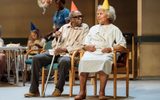 In the background is the remains of the party, there's a balloon with the number 80 on and an older lady eating cake. In the foreground Louis Mahoney (in a striped shirt and jeans but with a crutch in an orange party hat) and Cleo Sylvestre (in a hospital gown and a yellow party hat). Cleo is smiling and Louis is looking at her.