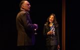 In the foreground of the photo is an out of focus man (Robert Glenister) who is looking outwards as if towards the audience. In focus further back in the photo is a woman with long dark curls (Vineeta Rishi). She's wearing a dark suit and a colorful scarf tied tight at the neck. She's holding a glass of wine.