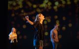 The main focus of the photo is Leah Gayer who is centre stage. The photo captures her from the side, her arms are flung open, holding a glass in one. Her long blonde hair is behind her ears and she is wearing blue denim shorts and a smart black top that looks velvet. In the background we see a man and a woman who are out of focus and projected on the screen behind looks like fairy lights.