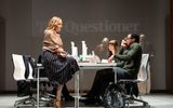 An office scene on the stage. On the screen at the back is projected the words The Questioner in a newspaper style font. Joanne Froggatt is sat on one of the desks in the striped skirt with a paisley patterned scarf wrapped around her. Simon Manyonda is sat at one of the desks on the phone and looking afraid.