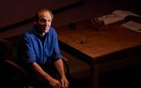 Taken from a higher angle looking down on the stage. We see a man (Ralph Fiennes) sitting on a chair next to a large wooden desk. On the desk are open books, glasses, glasses case, a glass jug. Ralph is wearing a blue shirt with the long sleeves folded up to his elbow. His hands are together in his lap and he looks lost.