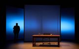 A low lit stage. At the back are 3 large square screens which take up the back of the stage. The one in the middle overlaps the two others. They are illuminated in a blue colour. To the left we see the silhouette of a man holding a bag on his shoulder. In front of the screens is a large wooden desk with a chair behind it. On the desk is a glass jug of water, a glass and a pot of pens.