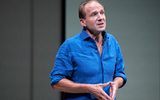 Close up of a man (Ralph Fiennes) in his 50's. He's wearing a blue shirt with the sleeves folded up to the elbows. His hands are together at his stomach, thumbs and fingers touching together gently.