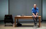 A large wooden desk with a chair next to it. On the chair is a man's blazer. There's a bag by one of the table legs. In the background are plain screens currently lit to be a grey colour. A man (Ralph Fiennes) sits on top of the table, hands together between his legs and ankles crossed together. He looks deflated.