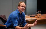 A man (Ralph Fiennes) sat in a chair next to a large wooden desk. The stage is well lit. Ralph is leaning forward with his elbows resting on his knees and his hands out as if explaining something. He's in a blue shirt with the sleeves rolled up to the elbows.