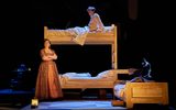 Lit on the stage is a wooden bunk bed and a small wooden bed which has a young boy sleeping in it. Samuel Blenkin is sat upright on the top bunk in a night shirt. Pandora Colin is leaning against the bunk bed frame with her arms crossed and looking off into the distance.