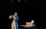 A chandelier hands above the scene. Samuel Blenkin is in a white curled wig and playing the harpsicord. Accompanying him is Pravessh Rana playing a wooden flute. He's wearing regal suit with knee high boots.