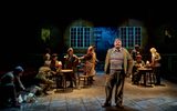 The cast of The Book of Dust on stage, there are wooden tables and chairs with cast sitting in a busy pub. Samuel Creasey stands centre stage.