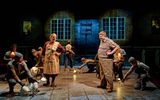 The cast of The Book of Dust on stage, there are wooden tables and chairs with cast sitting in a busy pub.