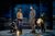 BoD 2-102 Left to right, Sky Yang (grey clothes and beanie hat) crouches with a puppet of a dog sniffing the ground. Ella Dacres (Brown shirt jacket, jeans) stands holding a rolled-up newspaper of chips. She looks at Pip Carter (tall in a long grey coat with a scarf), Julie Atherton (wears a full-body puppet of a hyena, she is bent over and laughing). In the background, the sign for the Trout Inn can be seen projected onto the back of the stage.