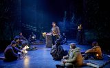 The cast of The Book of Dust sit scattered around the stage, looking up at John Light (wearing a flat cap and raincoat), he is next to a trash can fire.