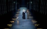 Two rows of wooden boxes line each side of the stage. Holly Atkins (dressed as a nun),holds Samuel Creasey (patterned jumper and plain khaki coat) by the ear. Left is Heather Forster crouching behind a box with a paper puppet of a cat facing the other two.