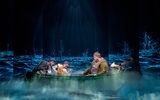 Ella Dacres (playing Alice Parslow) and Samuel Creasey (playing Malcolm Polstead), sit in a canoe shrouded in mist and lit by the lights in the paper puppets they hold. Projected onto the walls behind them is a dark forest. Sky Yang and Heather Forster crouch on either side of the canoe with paper puppets of a dog and cat placed on the canoe.