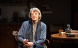 A woman, Maggie Smith, sits in a wooden chair next to a round wooden table. She has cropped grey hair and is wearing a long blue cardigan and blue tartan scarf draped down her. She's holding a pair of glasses in her hands and these rest on her crossed legs. She's looking up slightly and out into the audience. There's a spotlight on her and in the shadows in the background you can just make out the features of a kitchen.