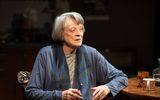 A close shot of a woman, Maggie Smith, sat at a round wooden kitchen table. She's got short grey hair and is wearing a long blue cardigan and blue scarf. She's looking to the side and holing a pair of glasses between her hands as if she's just taken them off and is closing them.