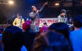 Production photo from Julius Caesar. Taken from the audience we see the tops of people's heads looking up to a stage with a band on, as if at a gig. The lead singer has the mic to his mouth and his left arm raised. Behind the band is a banner that reads 'Do this' in red.