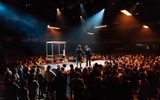 Production photo of Julius Caesar. A wide shot of the auditorium. In a raised platform amongst one hundred standing audience members Michelle Fairley and Adjoa Andoh shake hands.