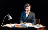 Production photo of Ben Whishaw in Julius Caesar. He's sat at a desk covered in books, wearing a black suit and glasses looking inquisitive and busy.