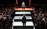 Production photo from Julius Caeser. Taken from high up on one end of the auditorium there are crowds of standing audience members either side of wide steps that take up the length of the room. At the top of the steps is David Calder as Julius Caesar sat in a red velvet chairs, either side of him are red flags flying. We see the back of someone who stands before him.