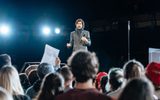 Production photo from Julius Caesar. Taken from the audience we see the back of peoples heads, someone is holding up a poster that we can't see. On a raised platform stands Ben Whishaw in a grey smart coat and stripey scarf holding a microphone like he's making a speech. He looks worried.