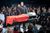 JC DR1-185 Production photo from Julius Caesar. On a slightly raised platform in the middle of crowds of audiences. David Morrissey stands on the platform above an open casket with David Calder lying dead as Caesar. Morrissey is holding his right hand up which is covered in blood.