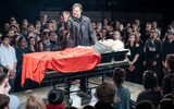 Production photo from Julius Caesar. On a slightly raised platform in the middle of crowds of audiences. David Morrissey stands on the platform above an open casket with David Calder lying dead as Caesar. Morrissey is holding his right hand up which is covered in blood.