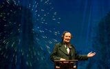 Photo from the production of A Very Very Very Dark Matter. Jim Broadbent is stood behind a wooden lectern that has an old book on it. He's wearing period clothing including waistcoat, smart jacket and patterned neckerchief. His hair is parted and curled on each side. His left arm is held out as if presenting something. Behind him is a projection of fireworks exploding in a night sky.