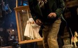 A close shot of the stage, in portrait angle. A full length Jim Broadbent stands leaning forward on one leg as if towards the audience, confiding in them. He's wearing pale brown subtly striped trousers, long smart black jacket and brown neckerchief. His hair is parted to one side and curled. The background looks like an attic and has wooden puppets hanging up around the place. Slightly obscured by the man we can see a glass and wooden box hanging from the ceiling with a girl leaning against the glass.