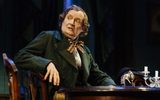 A close shot of Jim Broadbent sat at what would be a grand dark wooden table. He's looking back over his right shoulder with his right arm on the back of his chair, he looks surprised. He's wearing the brown neckerchief and his hair is parted and curled but grey.