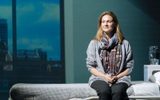 Production photograph from My Name is Lucy Barton. Laura Linney sits on a hospital bed with her hands together in her lap. She is wearing a purple scarf and smiling slightly. Behind her is a projection of a view of a city as if through the hospital window.