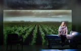 Production photograph from My Name is Lucy Barton. Laura Linney is sat on a hospital bed but the light shadows it as it's not a part of the scene. The back wall behind is filled with a projection of a corn field and an atmospheric sky above it.