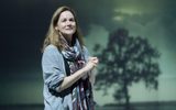 Production photograph from My Name is Lucy Barton. She is stood with her hands held together up at her chest, she's looking off into the distance wistfully. She's wearing a grey long cardigan and purple light scarf is wrapped around her neck. The silhouette of a tree is projected behind her.