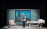 Production photograph from My Name is Lucy Barton. She stands centre stage with her hands together. There's a hospital bed to the right and a white hospital chair to the left. Behind is the large window frame filled with a projection of the view of New York, with the Empire State Building.