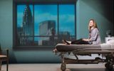 Production photograph from My Name is Lucy Barton. She is sat on the hospital bed with her legs out straight down the length of it. She looks out towards the camera. Behind is a large window frame filled with the projection of the view of New York.