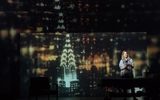 Production photograph from My Name is Lucy Barton. A darker image. Laura Linney is standing behind the hospital bed but it's shadowed out as not part of the scene. There's a single spotlight on her and the rest of the stage is flooded with a projection of a night time view of New York City. Taken from high up with a view of the Empire State Building, the lights of the city are what give the scene light.