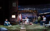 Nightfall production photo. An intricate set that looks like the garden of a house in the country. The stage is covered in grass with individual pavestones down the middle in a path. There's a picnic bench where Ophelia Lovibond (who has brown, mid-length hair, and is wearing a blue and white floral dress) is sat and Claire Skinner (who has cropped ash blonde hair, and is wearing dark blue jeans and long red cardigan) leans against. There is garden furniture about like a cushioned sun lounger. Ukweli Roach (wearing dark jeans, trainers and a polo top) sits on a chair looking down. Sion Daniel Young (wearing light blue jeans, white trainers and a blue polo top) sits on the grass with his arms on his knees. Everyone looks solemn. There's a wooden canopy covered in vines and fairy lights attached to a brick house at the rear of the stage.