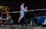 A garden scene, we can see the wooden picnic table and the edge of the vine and fairy lights hanging on the canopy. There's a huge copper pipe running across the back. Ophelia Lovibond is in a blue dotty dress and in the arms of Ukweli Roach. He's lifting her up as if she's run up to him or they're dancing.