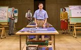 Samuel Barnett stands as the focus of the image in a blue shirt and brown smart trousers. He stands behind a big table with deign plans atop them. In the background are presentation boards with other architectural plans on.