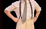 Ralph Fiennes stands with hands on his hips with his brows furrowed. He's wearing a white striped shirt, tie and braces.