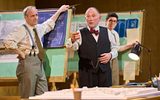 Ian Kirby stands leaning on a wooden board that shows blueprints and maps. He is wearing a white striped shirt, braces and a tie. Danny Webb stands with a cigar between his fingers talking. He is wearing a pin striped waistcoat and red silk bow tie. In the background Al Coppola is watching Danny Webb. He wears a white shirt and braces.