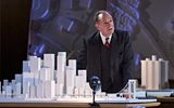 Ralph Fiennes looks pensive stood behind a 3D paper model of New York City, with sky scrappers and large buildings. He's wearing a dark suit and waistcoat and a red tie.