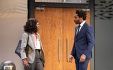 Lorna Brown and Yoli Fuller looking at eachother as if in confrontation, their brows are furrowed and they're both standing strong. Lorna has shoulder length curls and is wearing grey trousers and blazer with a leather hand bag over one shoulder. Yoli is in a navy suit with white shirt and maroon tie. In the background you can see the wooden double doors that lead to hotel corridor.