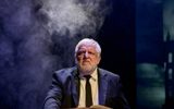 Simon Russell Beale sits behind a wooden chest wearing a suit and tie with a book in front of him and fog billowing behind.