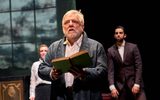 Simon Russell Beale stands with a book open in his hands looking upwards slightly.