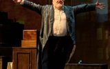 Simon Russell Beale stands with his arms outstretched wearing a long coat and white shirt.